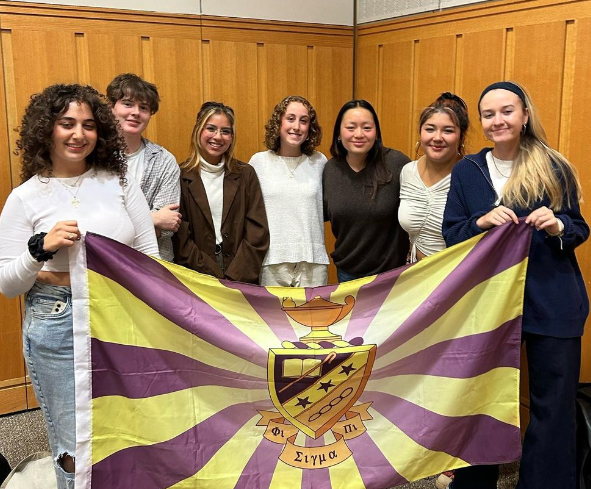 7 Phi Sigma Pi Members smile and hold a flag with the Phi Sigma Pi crest.
